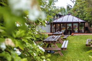Un porxo amb teulada metàl·lica, envoltat d'una zona verda amb taules de picnic.