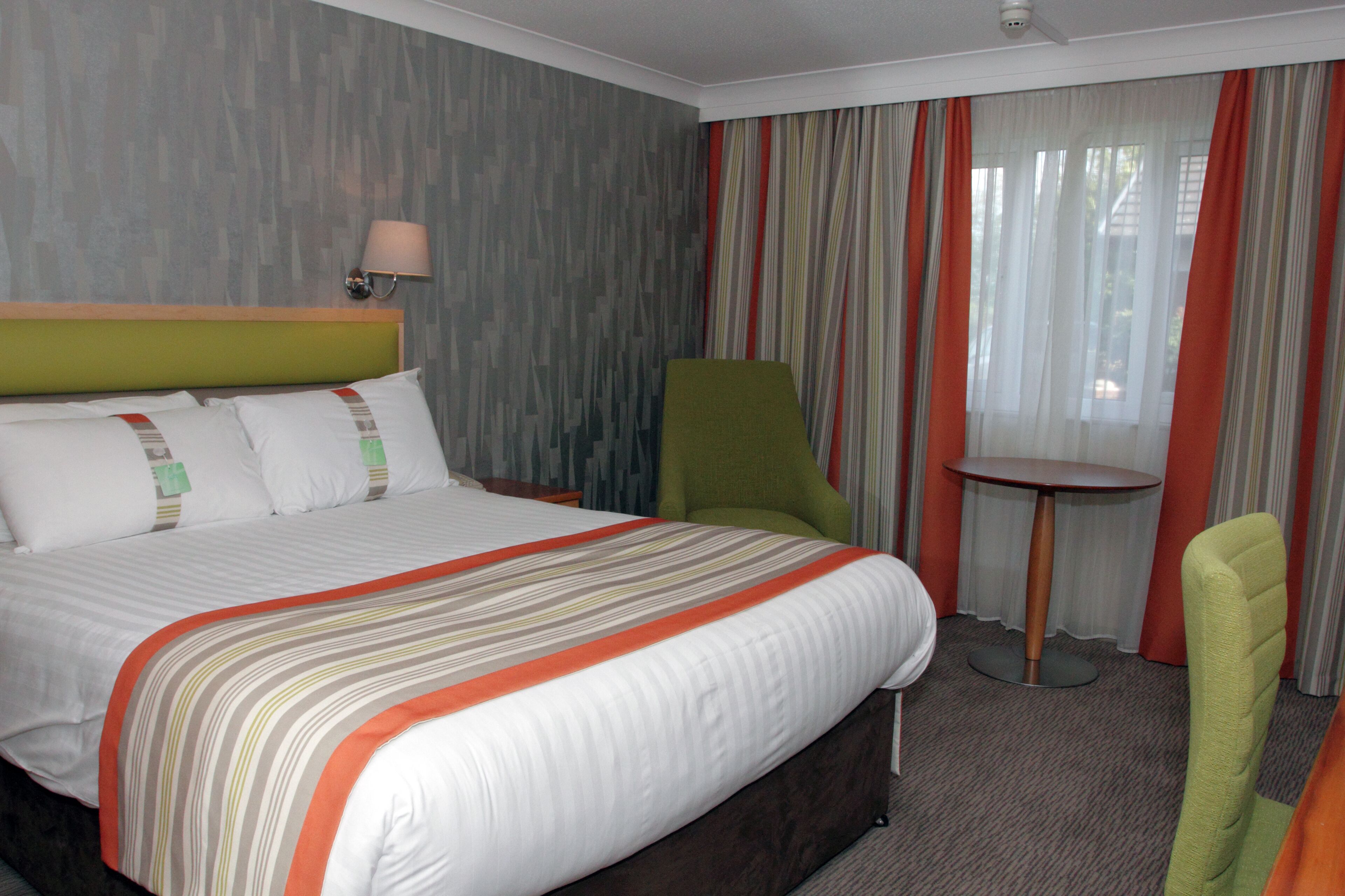 A hotel room with a large bed, a green chair, a small round table, and a lamp.
