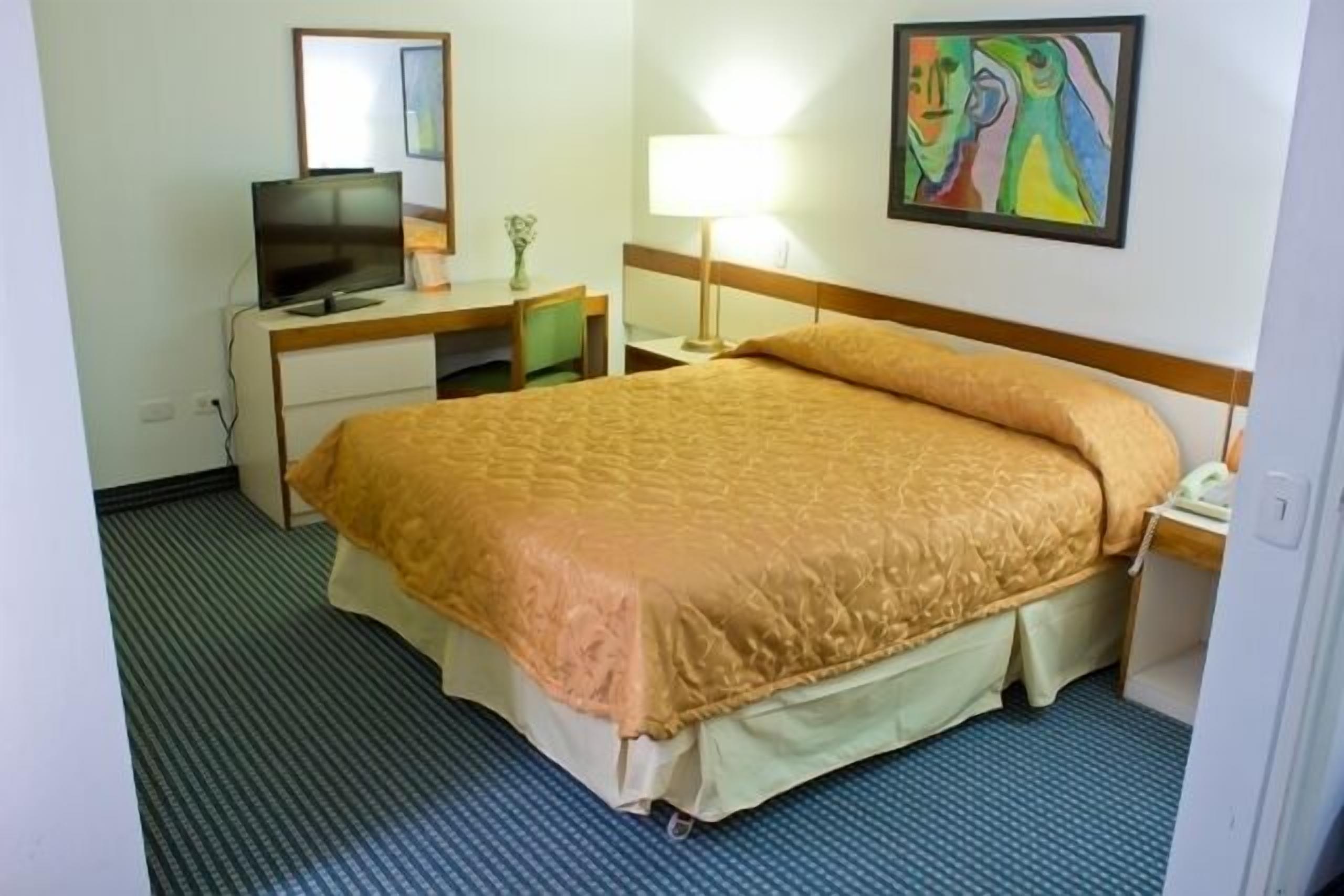 A hotel room with a bed, a desk with a computer, a TV, a lamp, and a painting on the wall.