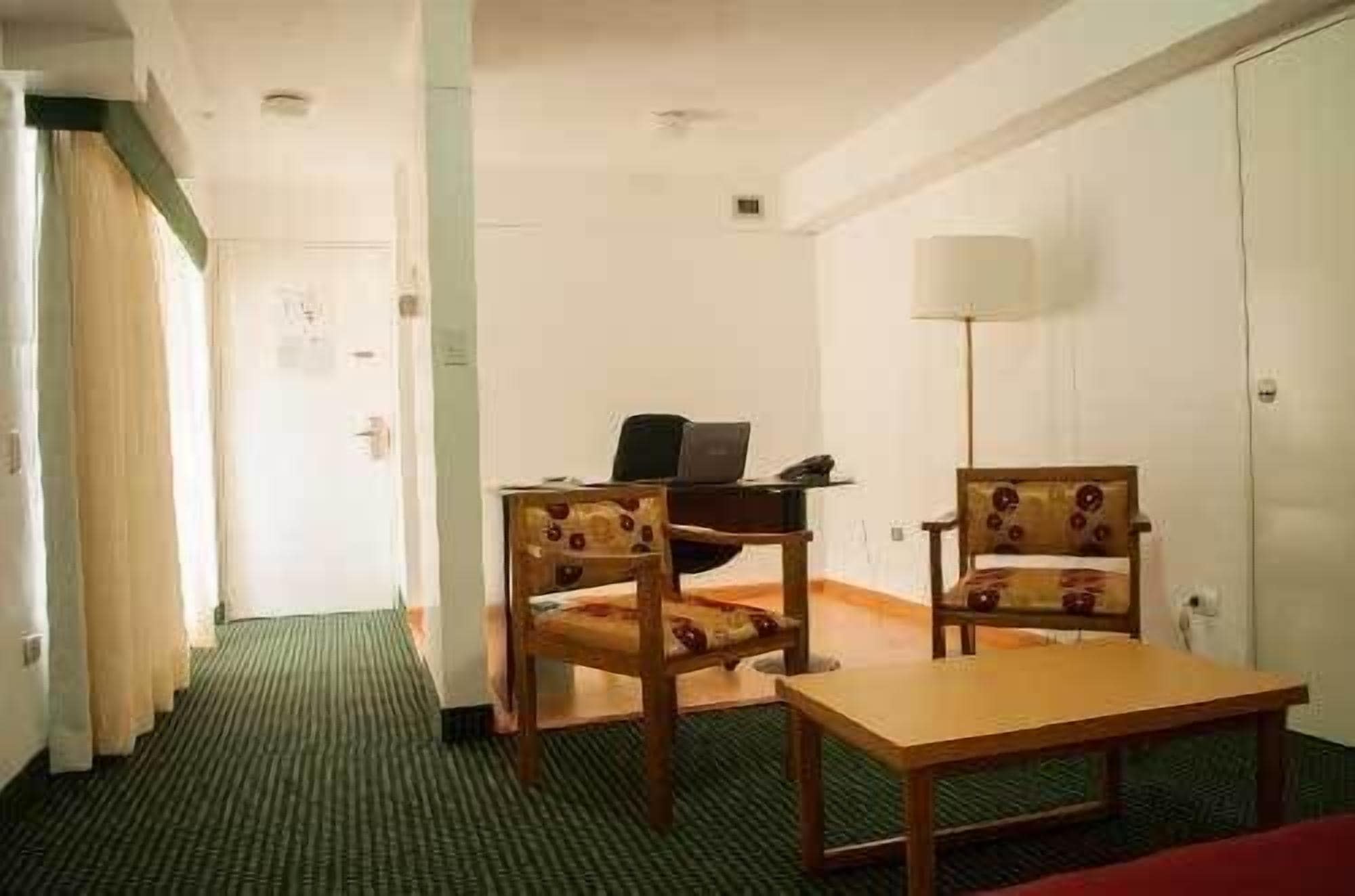 A hotel room with a desk, two chairs, a small table, a lamp, and a wardrobe.