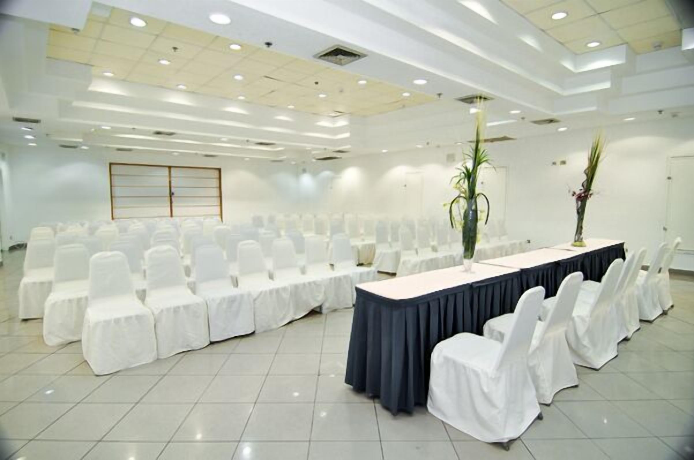 A conference room with rows of white chairs, a long table with a dark tablecloth, and two tall vases with plants.