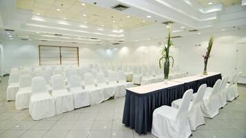 A conference room with rows of white chairs, a long table with a dark tablecloth, and two tall vases with plants.