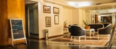 A modern hotel lobby with a reception desk, framed pictures on the wall, and a seating area with chairs and a table.