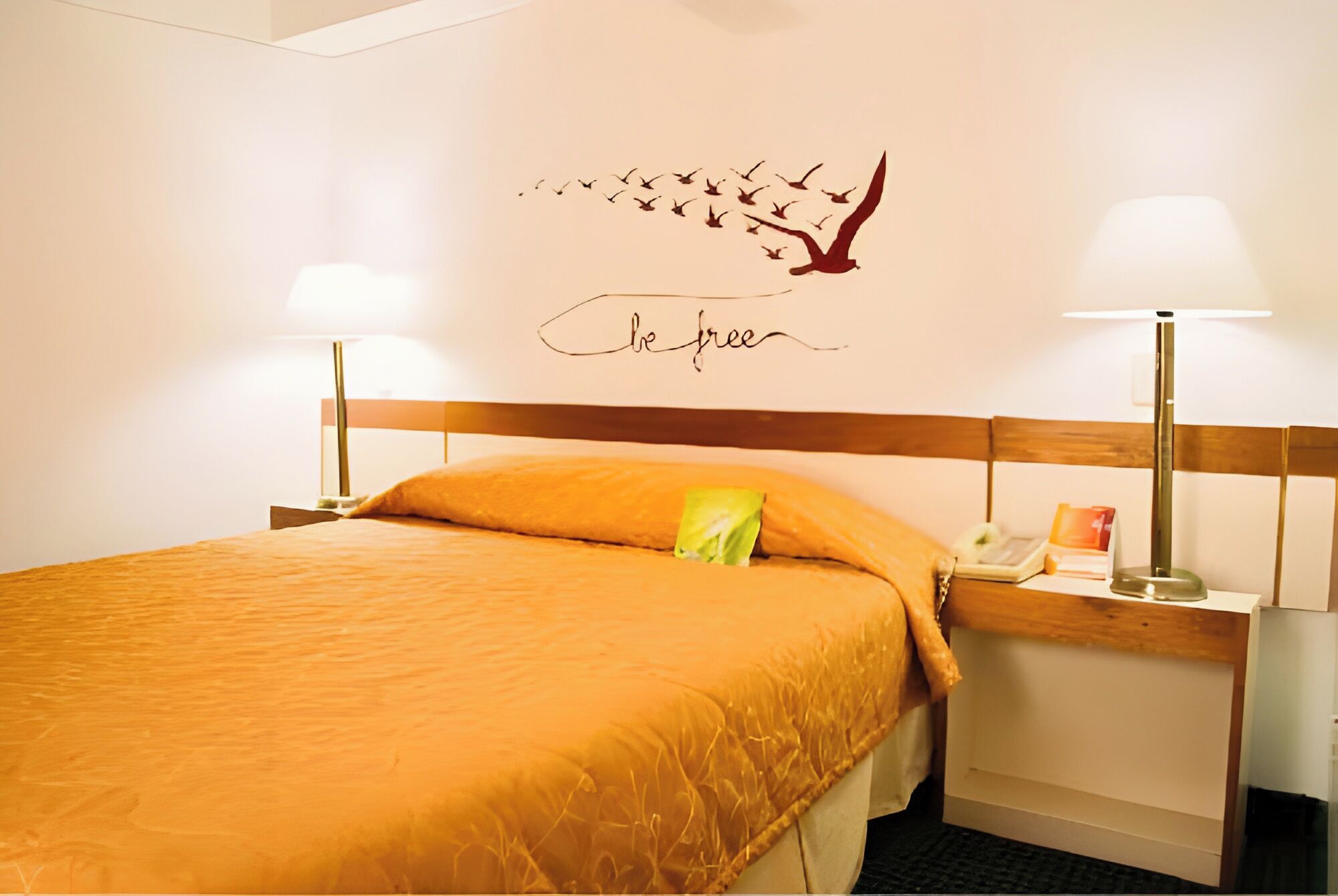 A neatly made bed with a wooden headboard, two bedside lamps, and a wall decoration of birds.