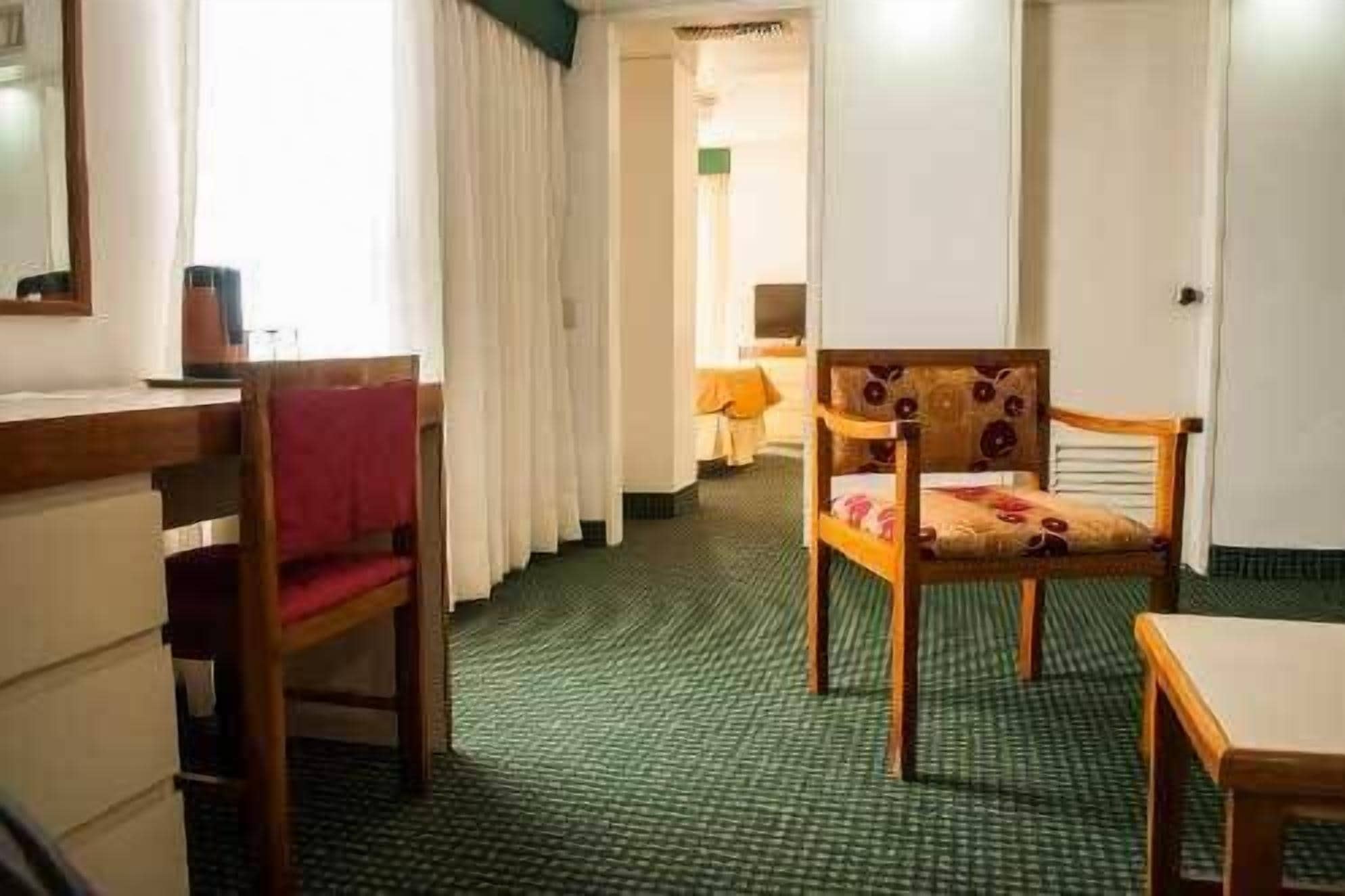 Ein Hotelzimmer mit grünem Teppich, einem hölzernen Stuhl mit floralem Kissen, einem Holztisch und einem Bett mit roter Decke.