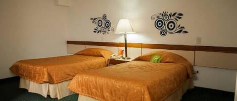 Habitación de hotel con dos camas, cada una con cubrecama naranja y falda blanca. Hay una mesita de noche con una lámpara y un teléfono.