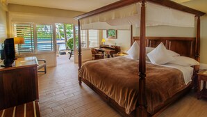 A four-poster bed with a brown bedspread, a desk with a lamp, and a chair in a hotel room with wooden floors and large windows.