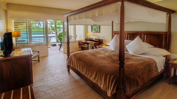A four-poster bed with a brown bedspread, a desk with a lamp, and a chair in a hotel room with wooden floors and large windows. -  Plantation Bay a Real Resort with a Real Spa (Lapu-Lapu)