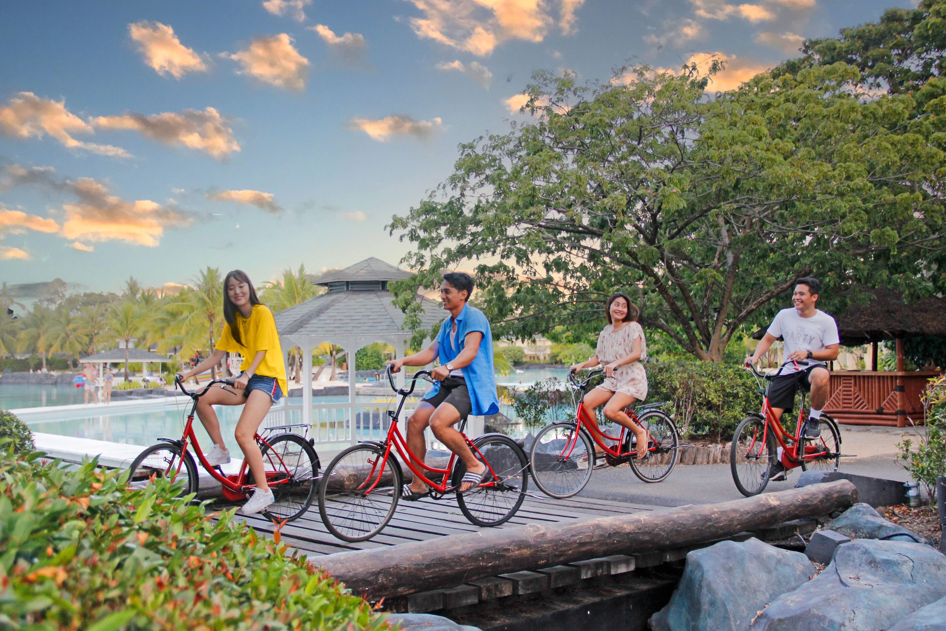 A group of people riding bicycles on a wooden bridge with a pool and trees in the background.