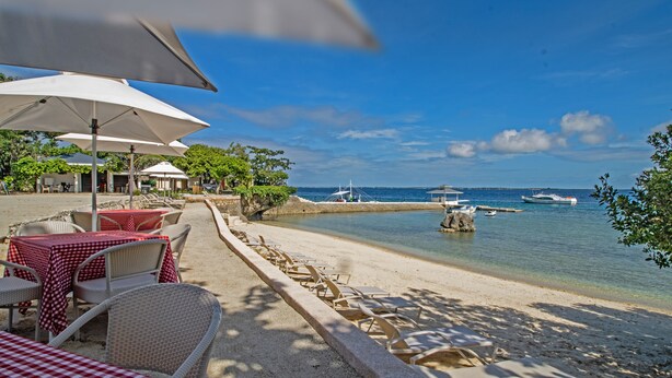 A beachside restaurant with outdoor seating, including tables and chairs, and a view of the sea. - Plantation Bay a Real Resort with a Real Spa (Lapu-Lapu)