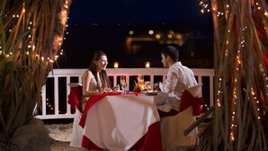 An outdoor dining setup with a table and chairs, surrounded by string lights and palm fronds.