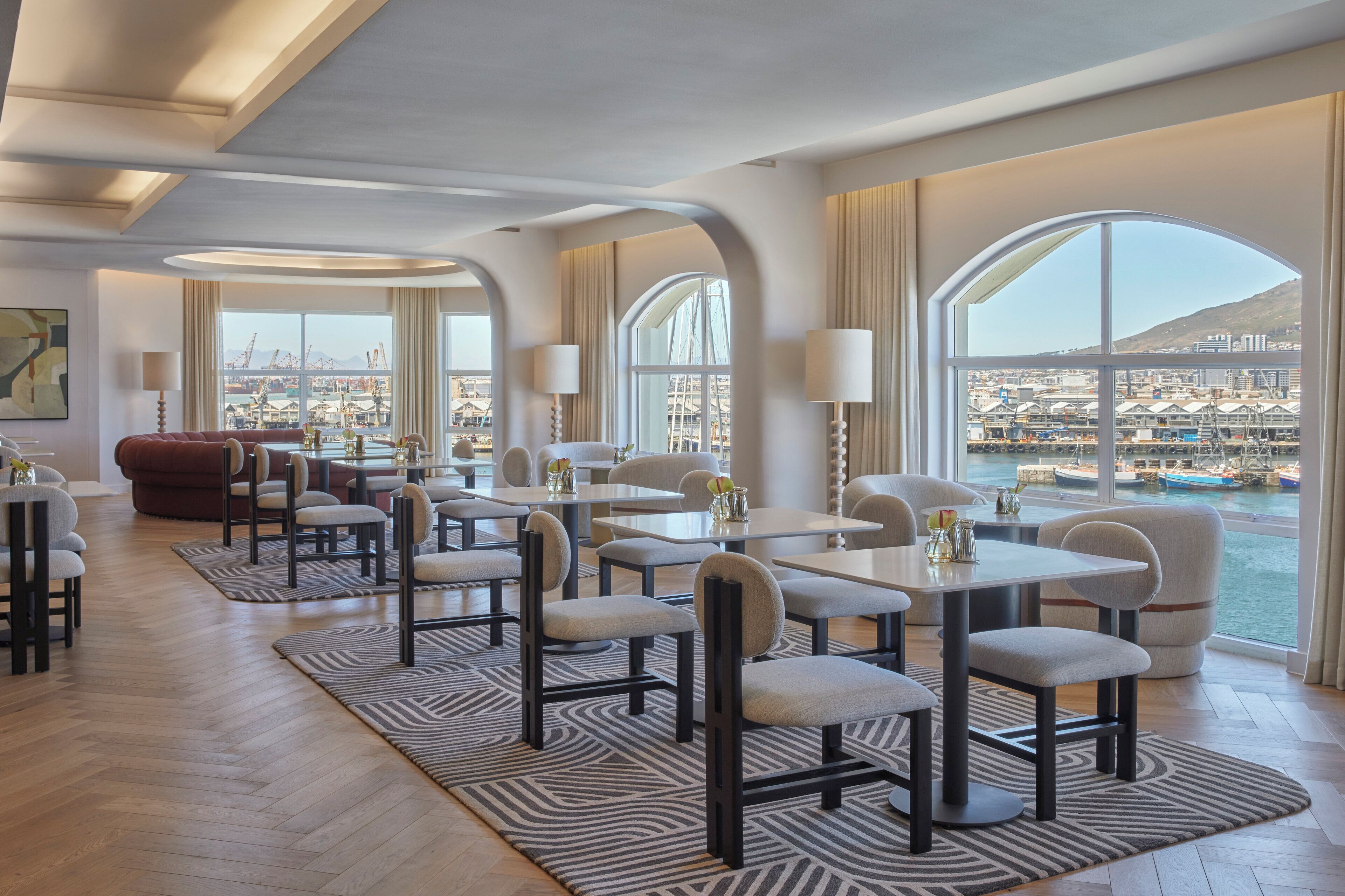 Un espace repas moderne, avec des tables et des chaises, de grandes fenêtres offrant une vue sur un port de plaisance et sur le paysage urbain, et un tapis à motifs sur le sol.