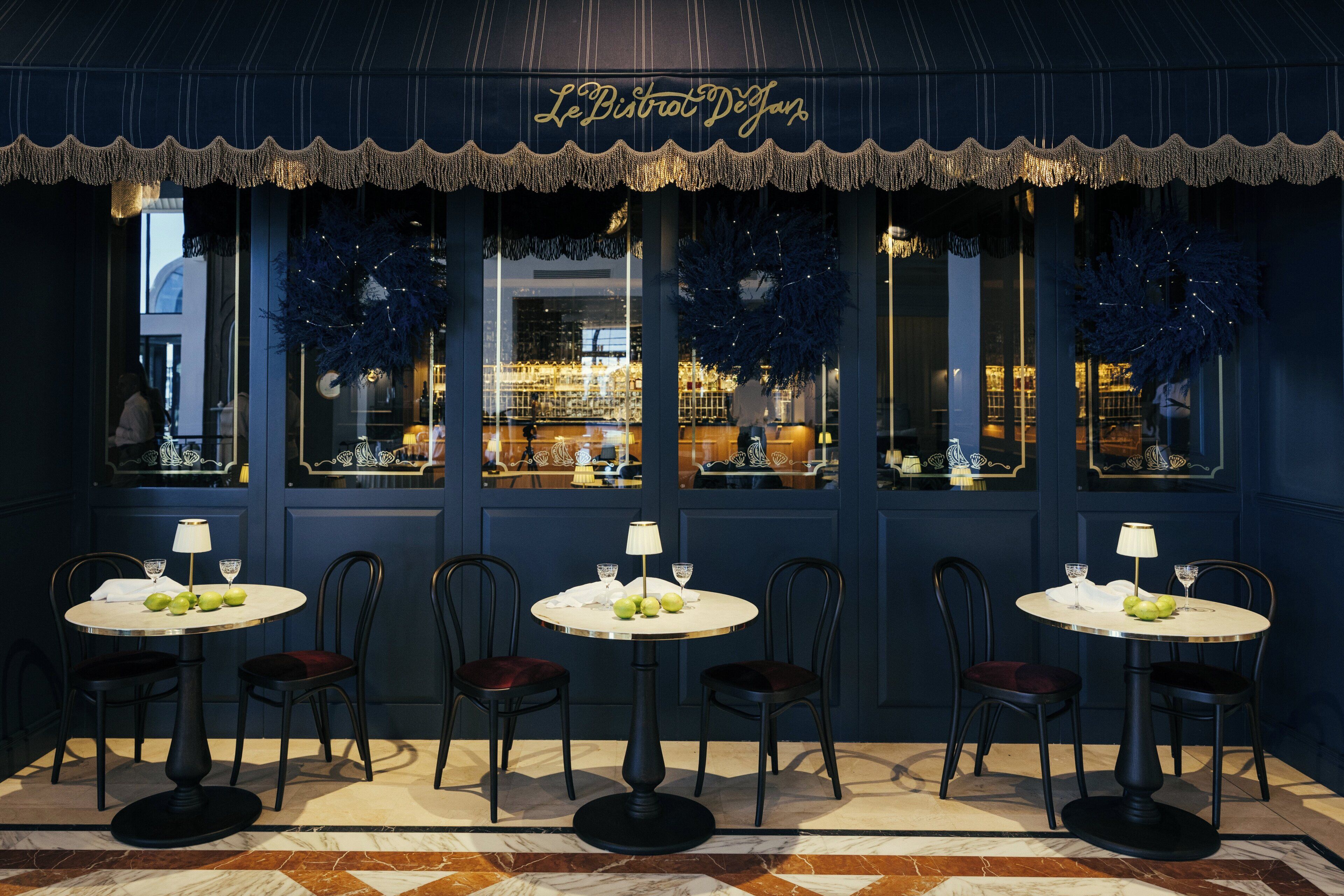 Un restaurante con fachada azul oscuro, cinco mesas con manteles blancos y sillas.