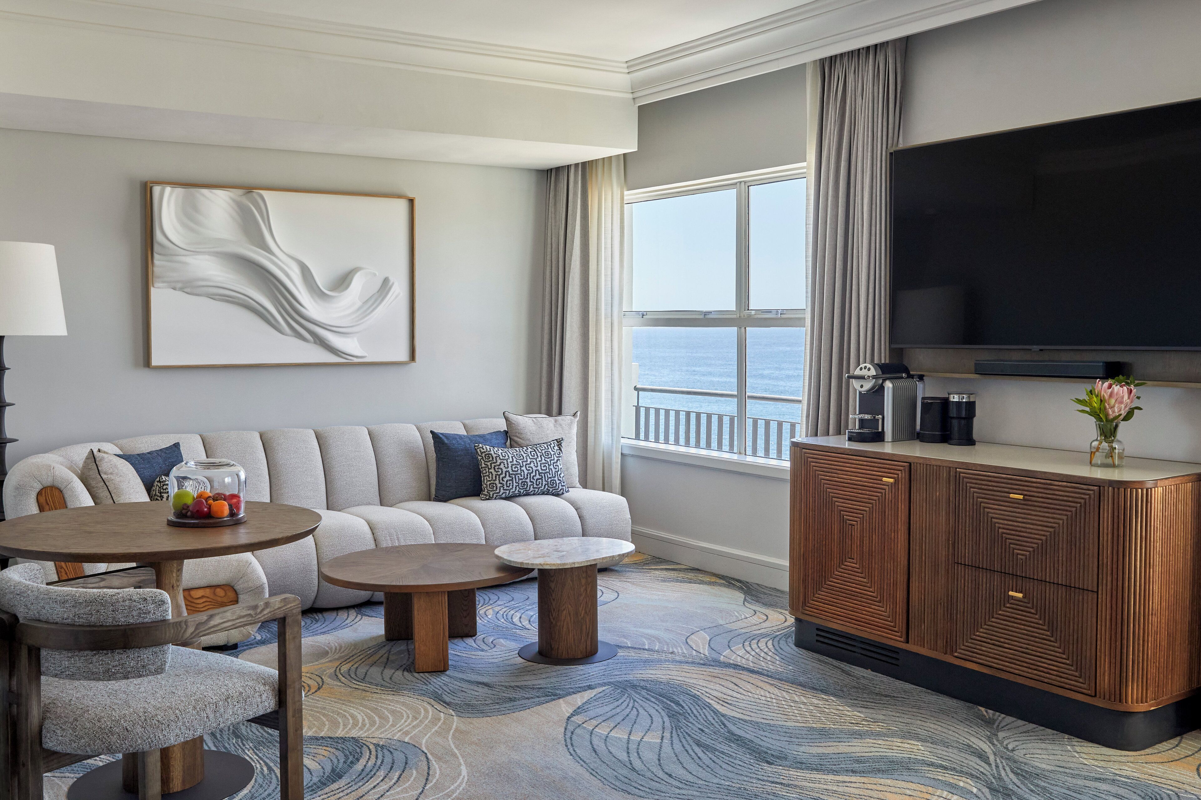 A modern living room with a sofa, two coffee tables, a TV, and a view of the sea.