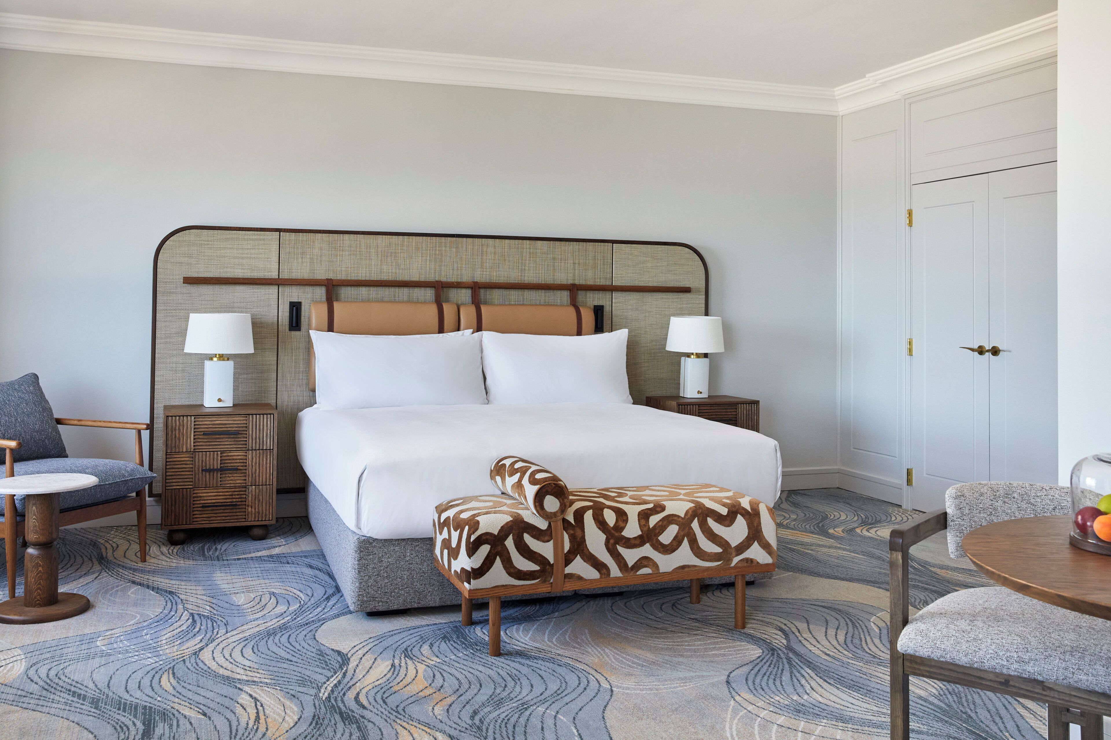 A hotel room with a large bed, two bedside tables, a chair, a small table, and a decorative wall panel.