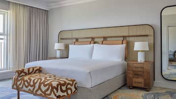 A neatly made bed with a headboard, two bedside tables with lamps, a patterned ottoman, and a large mirror.