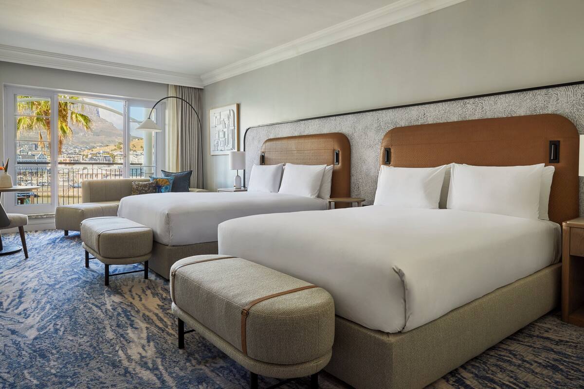 A hotel room with two beds, a sofa, a chair, and a view of a cityscape through large windows.