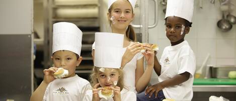 A group of children in chef hats and aprons, eating pastries in a kitchen setting.