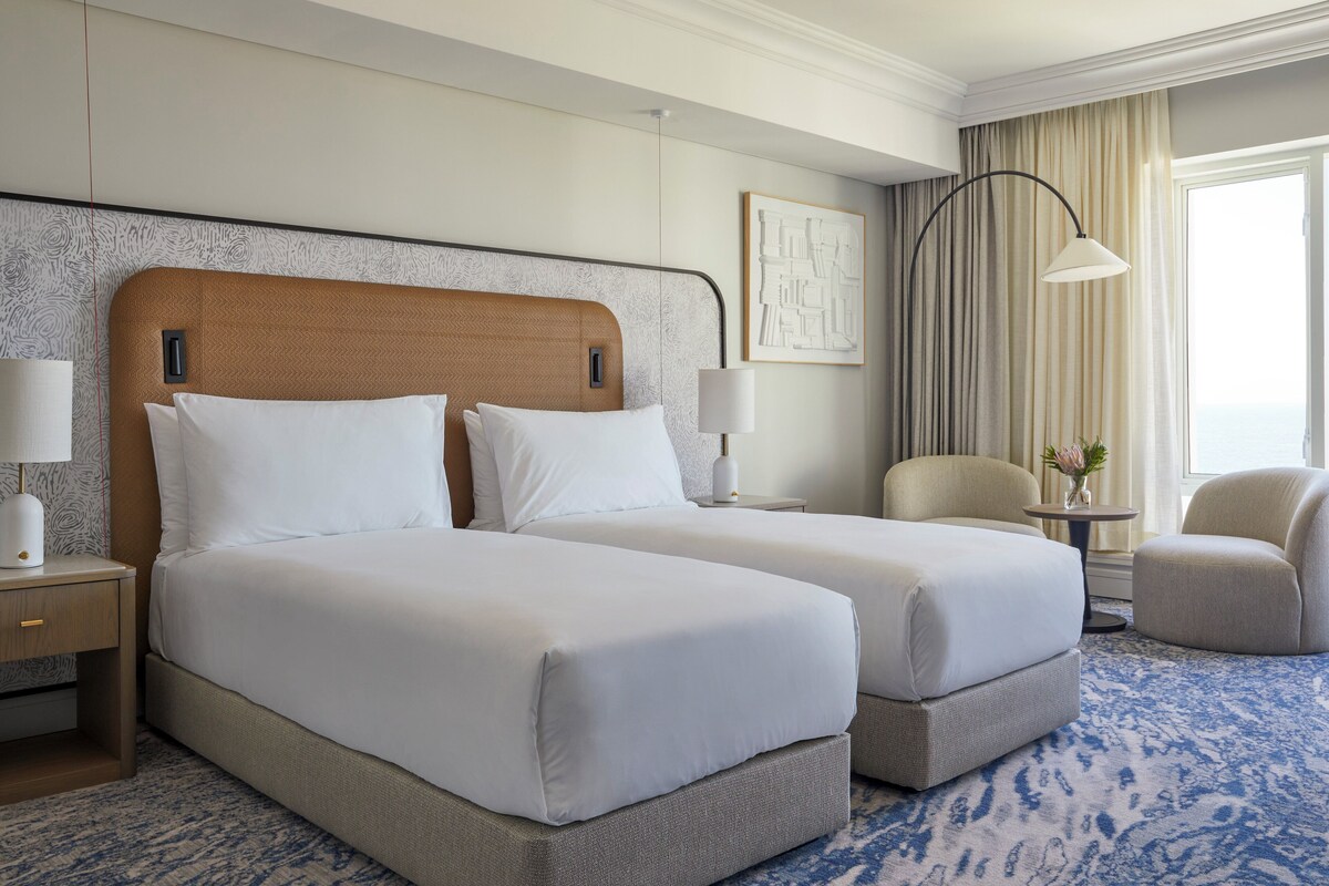 Une chambre d’hôtel avec deux lits, une table de chevet, une chaise, une lampe et une vue sur l’extérieur.