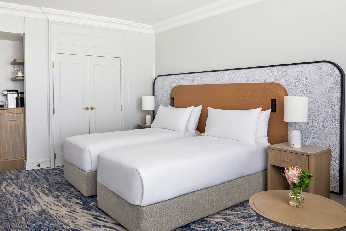 A double bed with white linens, a round wooden nightstand with a vase of flowers, and a white door with gold handles.