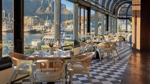 A restaurant with a checkered floor, tables set with white tablecloths, wicker chairs, and a view of a harbor with boats and buildings. - InterContinental Table Bay Cape Town by IHG (Cape Town)