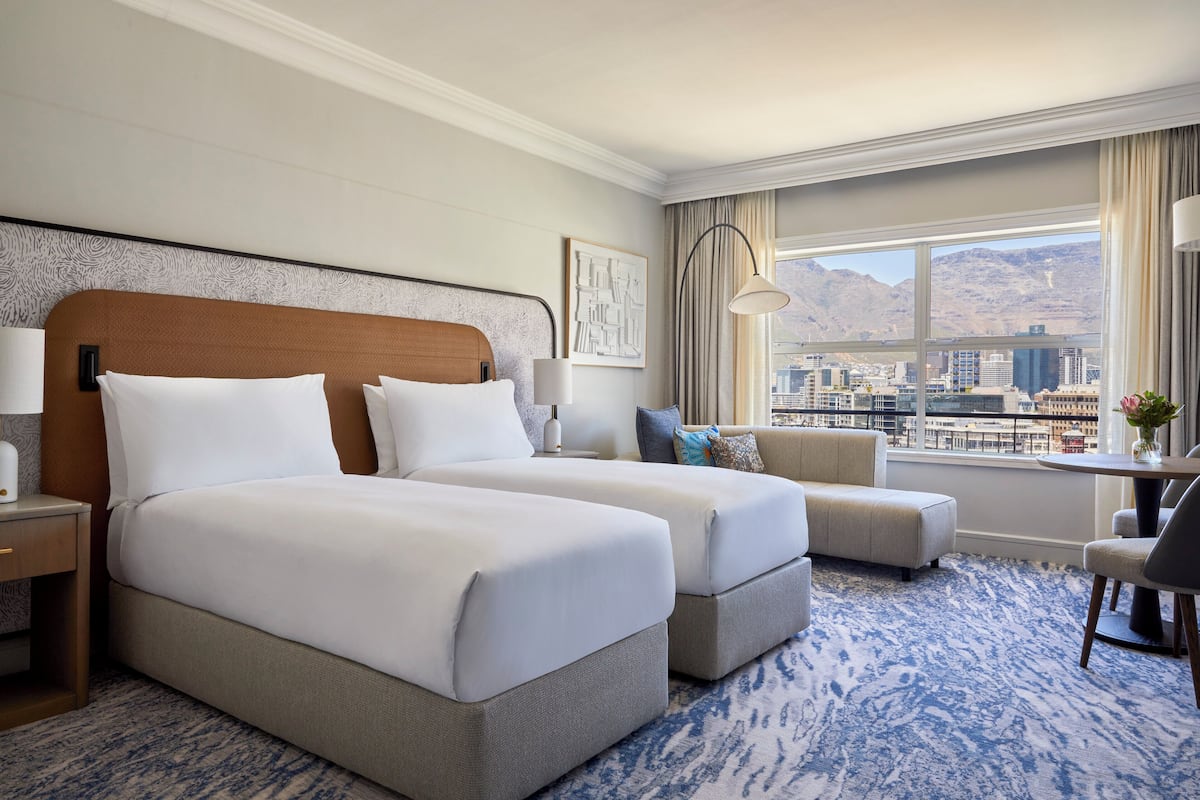 A hotel room with two beds, a sofa, a desk, and a view of the city and mountains.