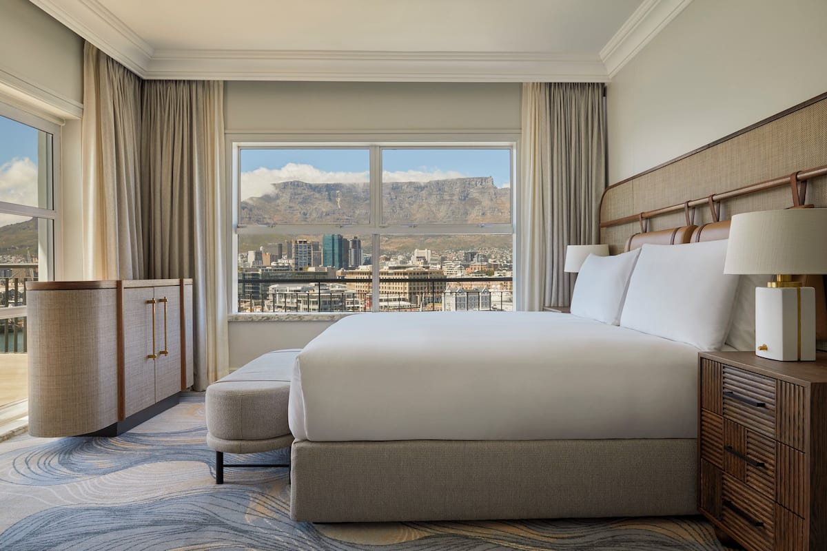 A hotel room with a large bed, a bedside table with lamps, a view of a cityscape, and a window with curtains.