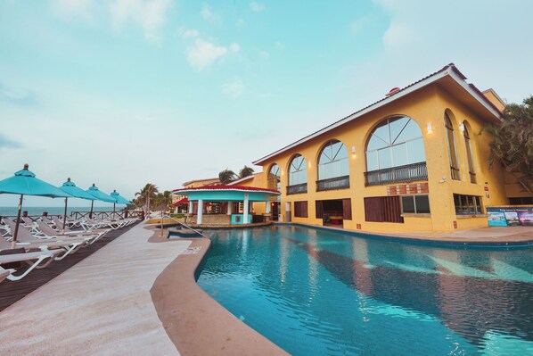 A poolside area with lounge chairs and umbrellas, a building with arched windows, and a clear sky.