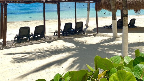 A beach with a thatched-roof structure, several lounge chairs, and palm trees. - All Ritmo Cancun Resort & Water Park - All Inclusive (Cancun)