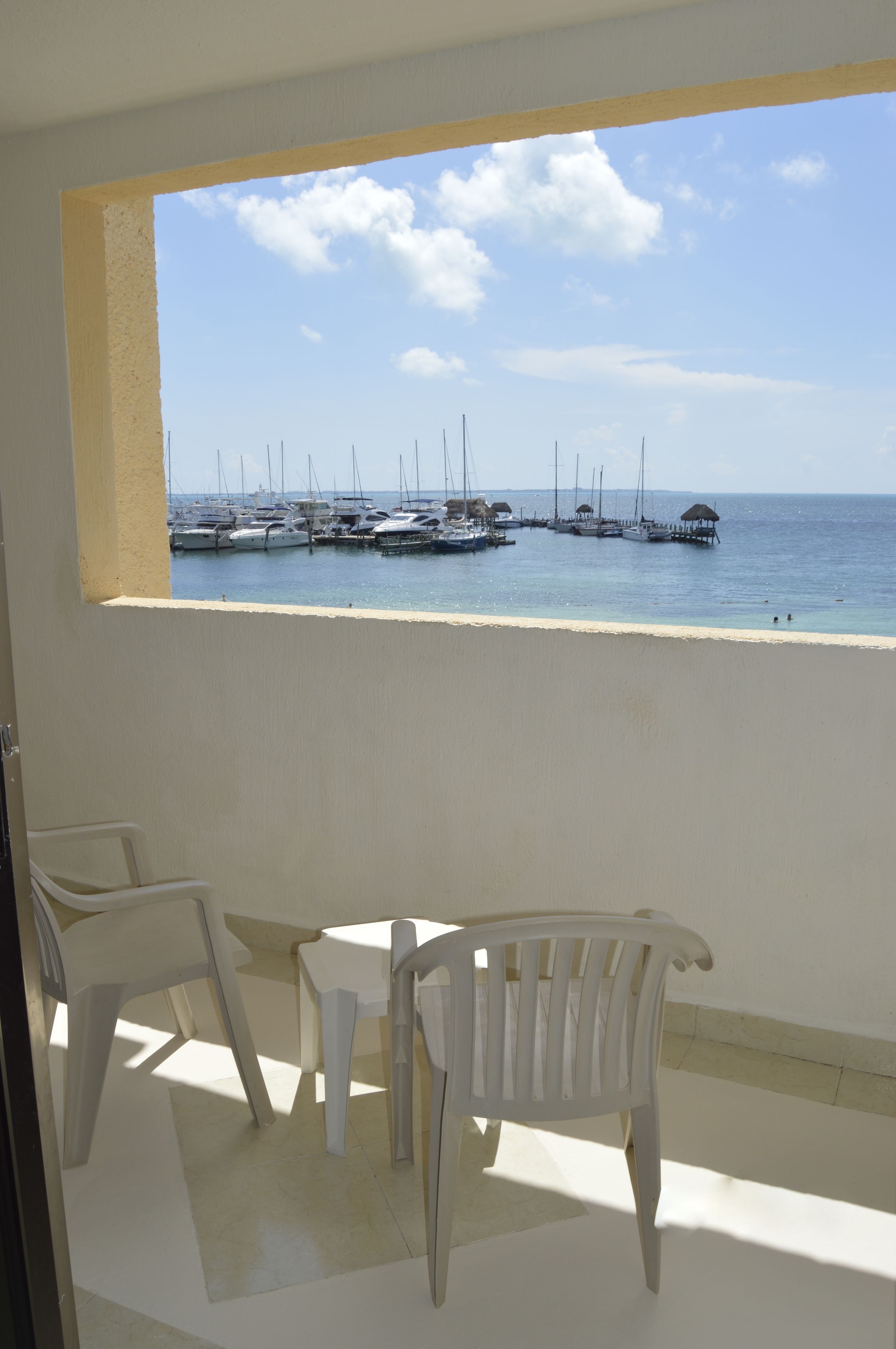 A balcony with white plastic chairs and a small table, overlooking a marina with several boats docked near the shore.