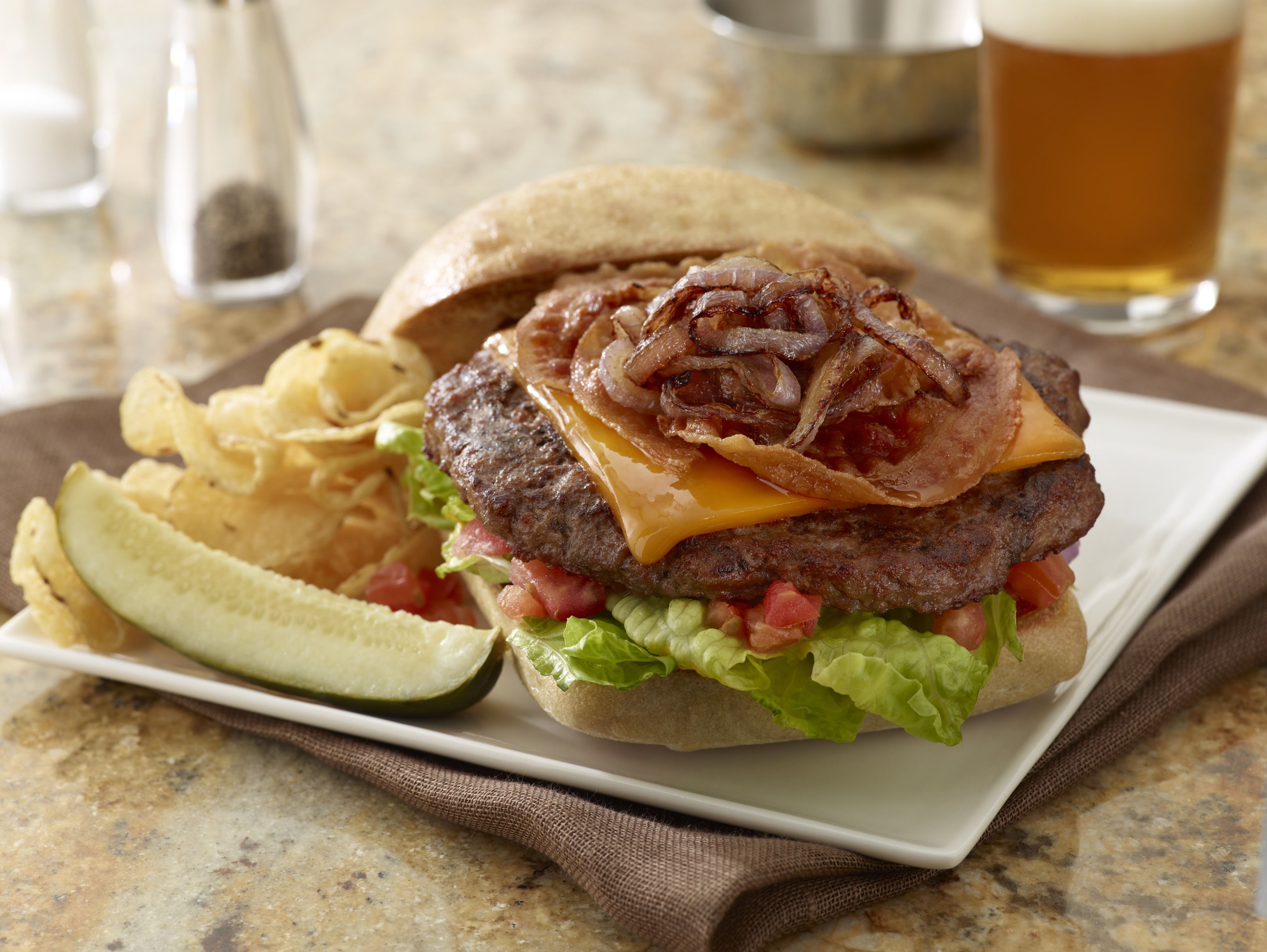 Hamburguesa con tocino, queso, lechuga y tomate en un pan, acompañada de papas fritas y pepinillo.