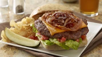 Hamburguesa con tocino, queso, lechuga y tomate en un pan, acompañada de papas fritas y pepinillo.