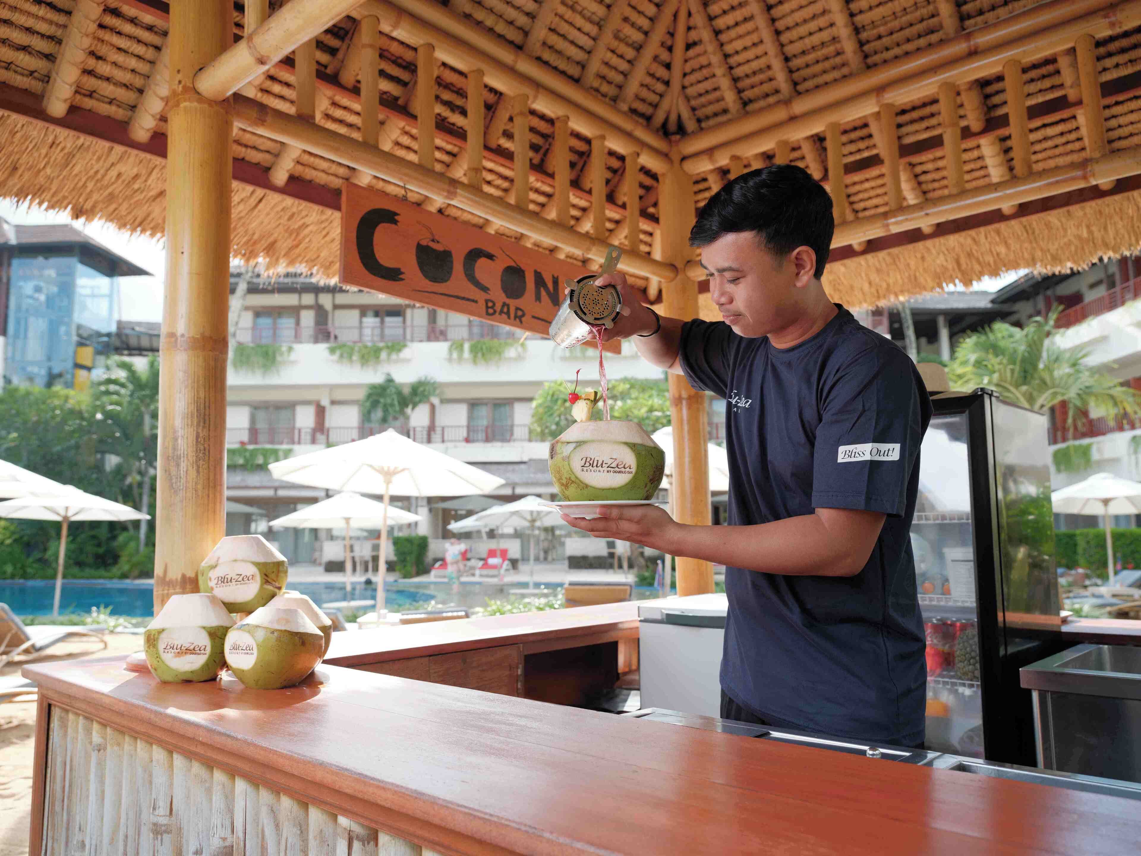Uma pessoa servindo bebida em um coco em um bar com uma placa indicando 'Coconuts Bar'.