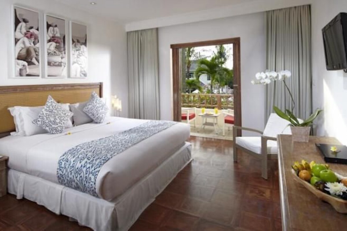 A hotel room with a large bed, a desk with a fruit bowl, a TV, and a balcony with a view of palm trees.