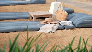 A beach lounge area with blue cushioned loungers, a wooden table, a woven beach bag, and a straw hat. - Blu-Zea Resort by Double-Six (Seminyak)