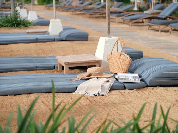 A beach lounge area with blue cushioned loungers, a wooden table, a woven beach bag, and a straw hat. - Blu-Zea Resort by Double-Six (Seminyak)
