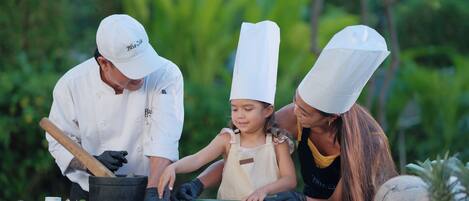 Un chef y dos personas preparando comida al aire libre, con una variedad de ingredientes frescos sobre la mesa.