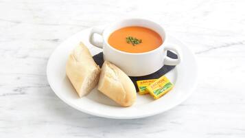 A white cup of soup with a garnish, accompanied by two baguettes and two packets of anchovies on a white plate.