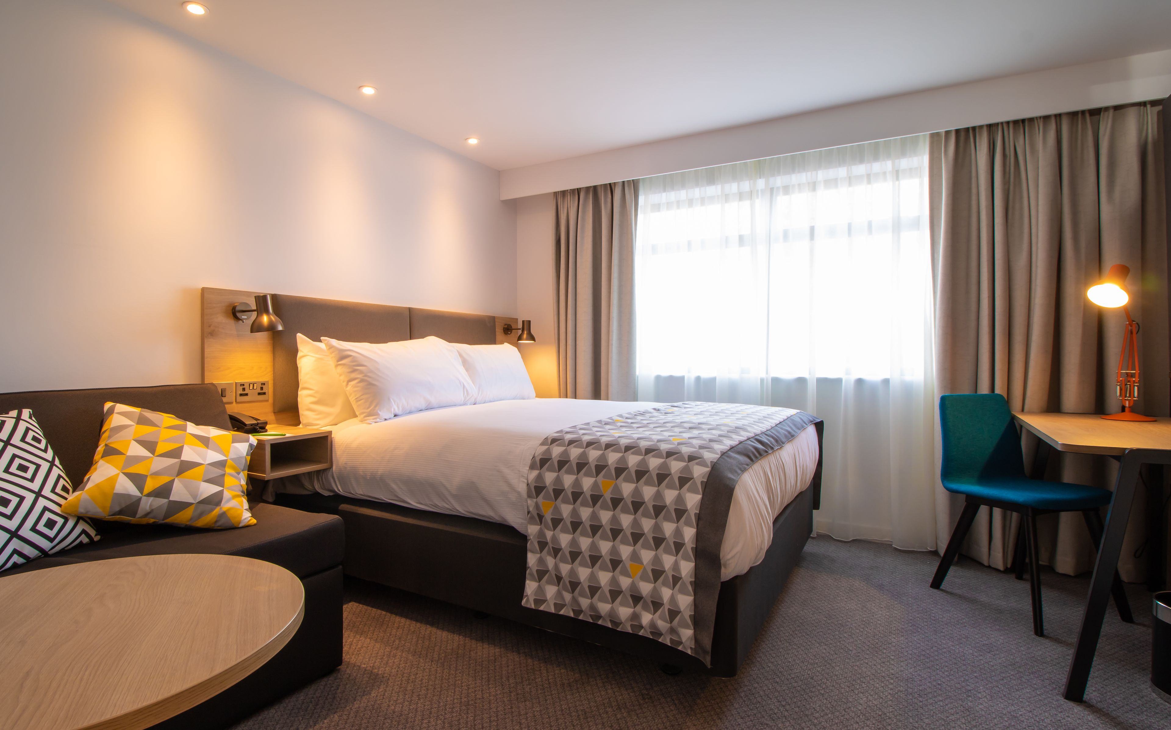 A hotel room with a bed, a desk, a chair, a sofa, and a lamp.