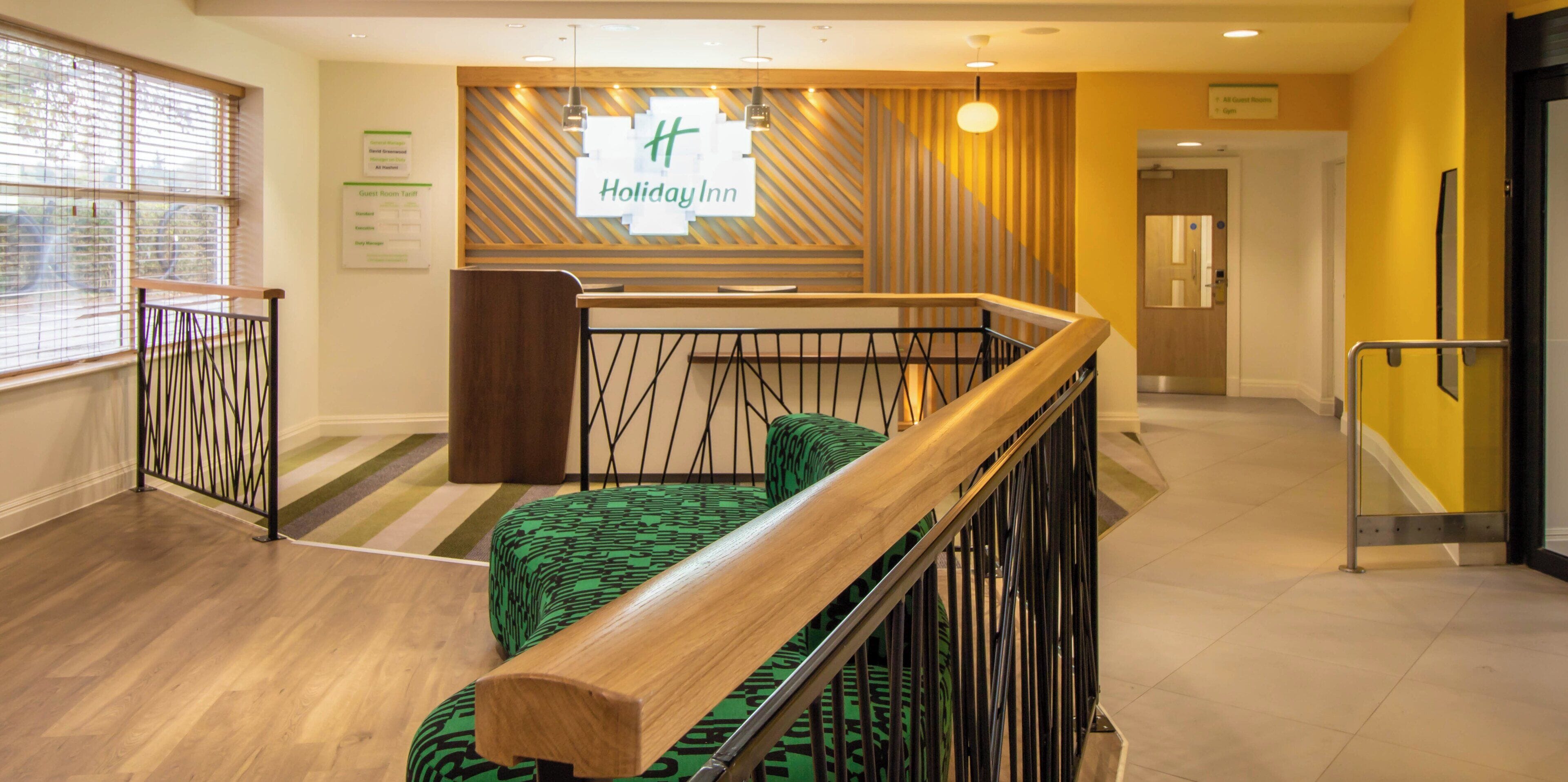 Un hall de hotel con mostrador de recepción, una escalera con barandilla de madera y un sofá con estampado verde.