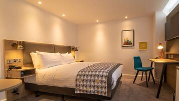 A hotel room with a bed, bedside table, desk, chair, and a wall-mounted TV.