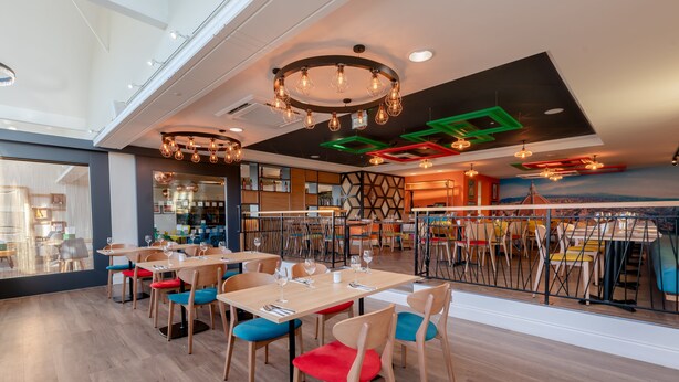 A modern restaurant with wooden tables and chairs, a high ceiling with exposed beams, and a view of the city through large windows. - Holiday Inn Leicester - Wigston by IHG (Wigston)