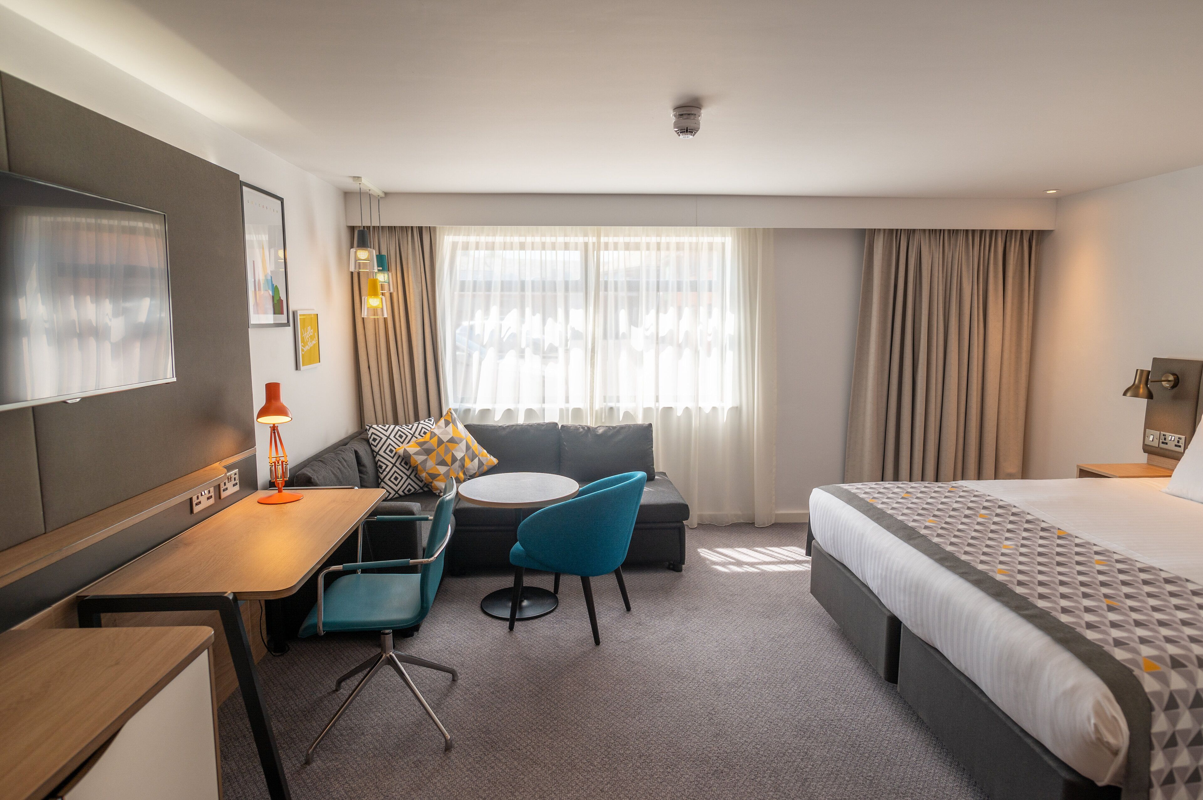 A hotel room with a bed, a desk, a chair, a sofa, and a window with curtains.