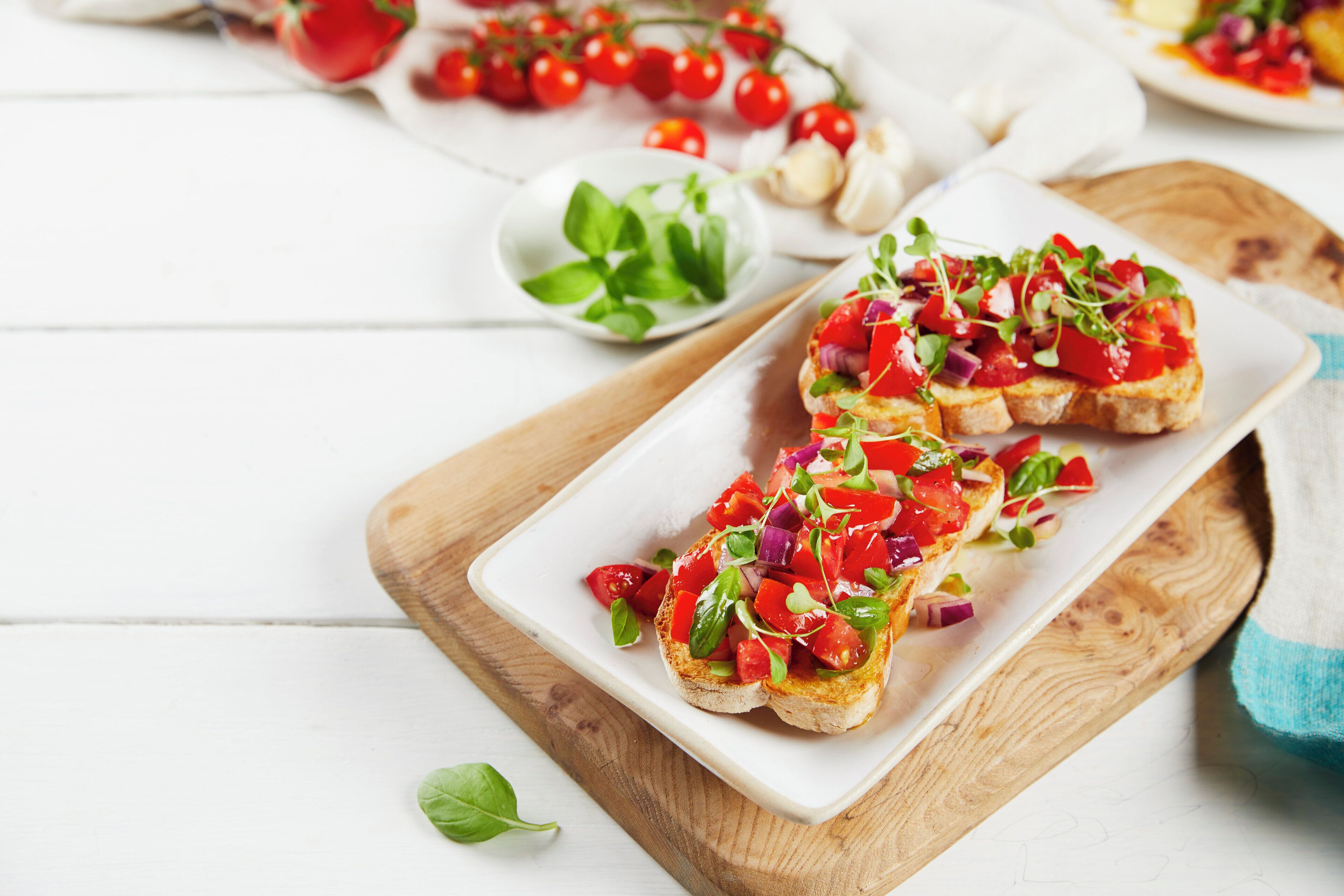 A wooden cutting board with a white plate containing a sandwich topped with tomatoes and greens.