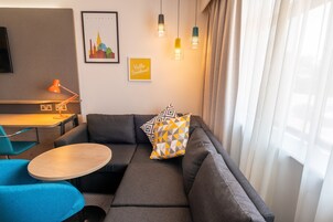 A modern living room with a sectional sofa, a small round table, a desk with a lamp, and a cityscape artwork on the wall.