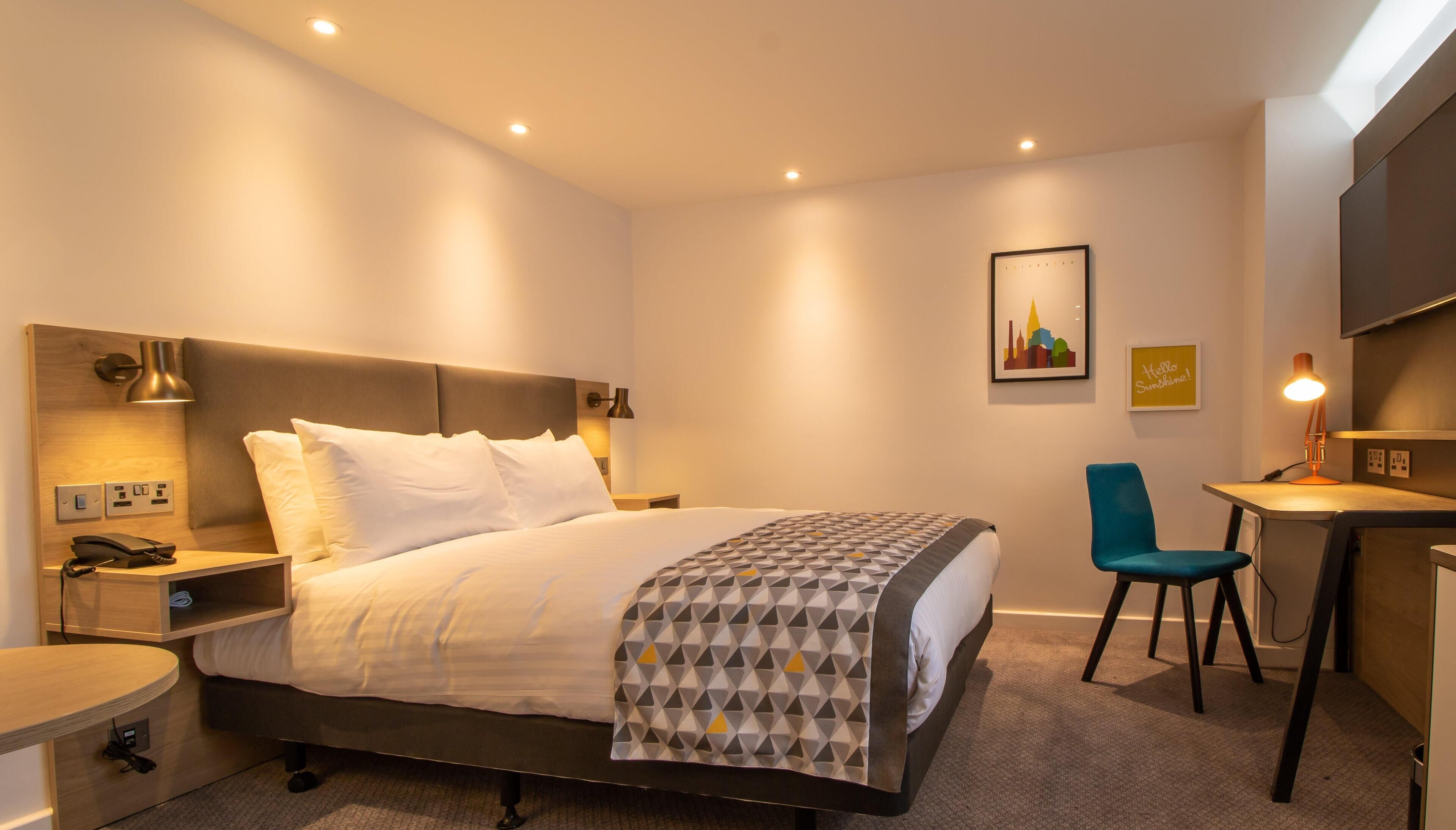 Ein modernes Hotelzimmer mit einem großen Bett, einem Nachttisch mit Telefon, einem Schreibtisch mit Stuhl und einem an der Wand befestigten Fernseher.