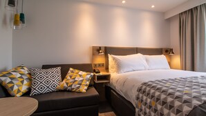 A modern hotel room with a bed, a sofa, and a small table. There are two bedside lamps, a nightstand, and a power outlet. - Holiday Inn Leicester - Wigston by IHG (Wigston)