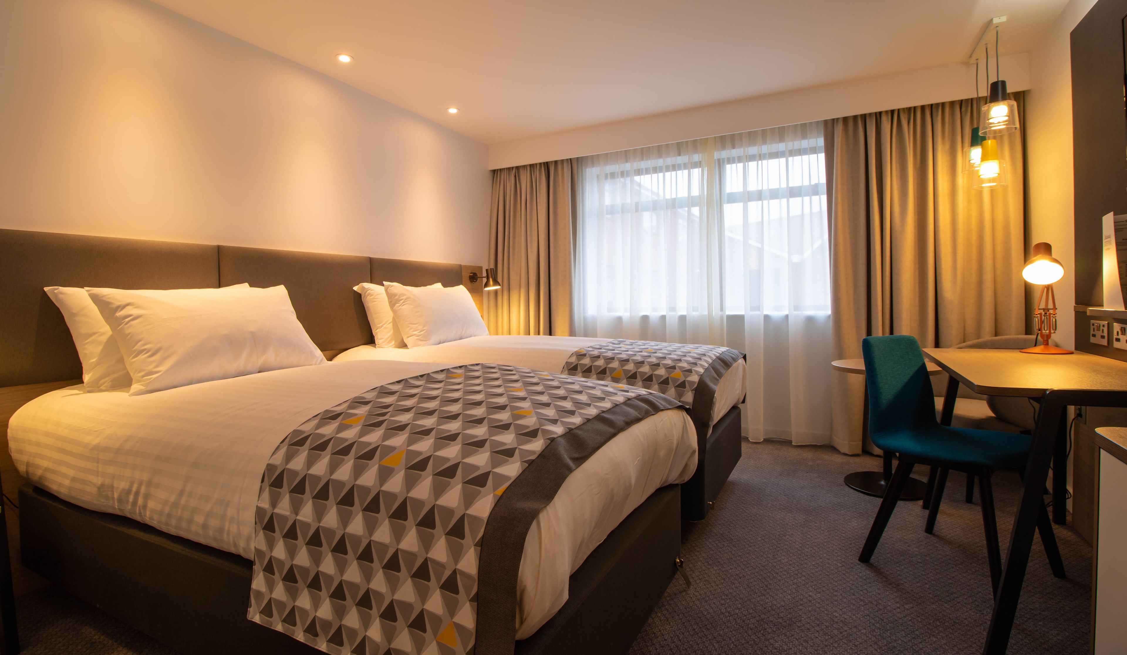 A hotel room with two beds, a desk, a chair, and a lamp.