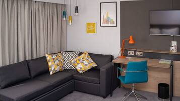 A modern hotel room with a sofa, a desk, a chair, a TV, and a curtain.