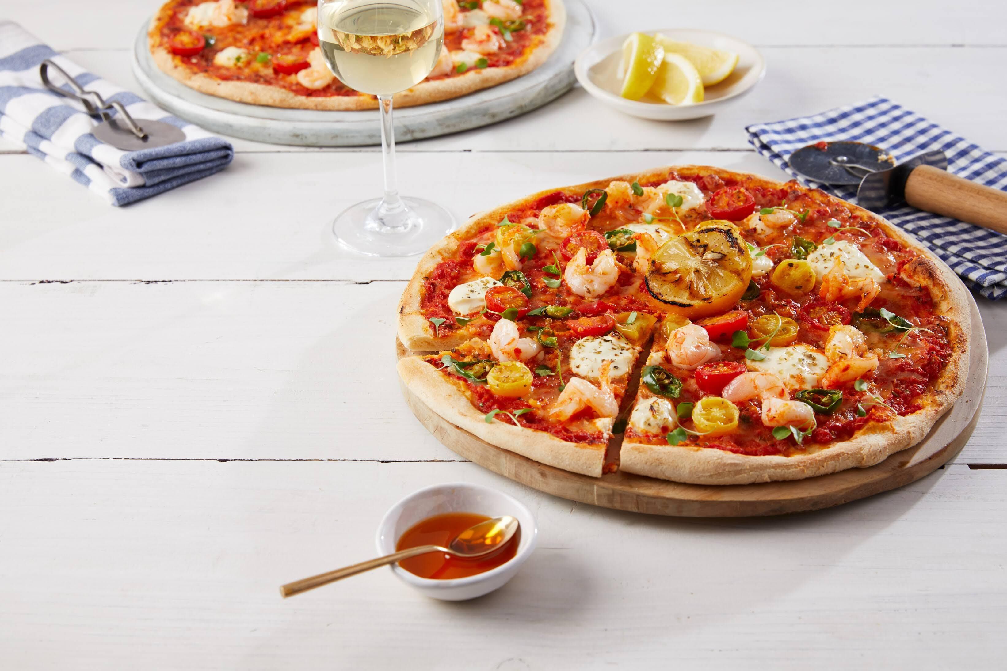 Zwei Pizzen auf Holzbrettern mit einem Glas Weißwein, einer halben Zitrone und einer kleinen Schale Soße auf einem weißen Holztisch.